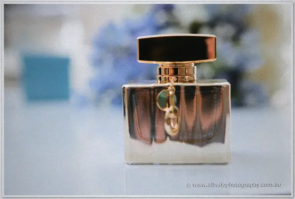 A perfume bottle with gold cap and charm waits on a white table, blue flowers soft behind