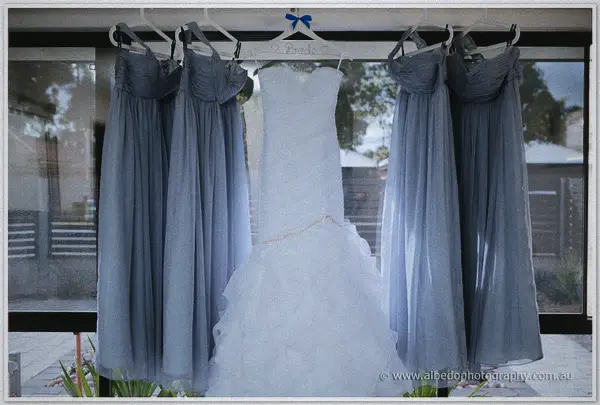 Sarah's ruffled gown hangs on a hanger flanked by four grey-blue chiffon bridesmaid dresses by the window