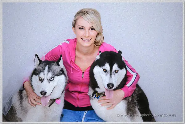Veiled Sarah in her pink hoodie cuddles her two huskies against a plain white studio wall