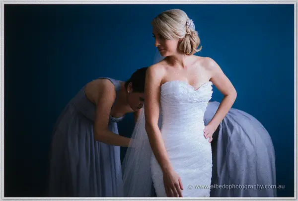 Sarah stands in her strapless lace gown against a deep blue wall as a bridesmaid kneels to arrange her veil