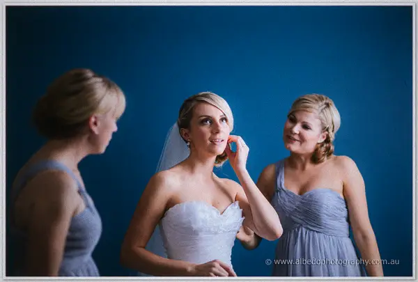 Sarah laughs between two grey-gowned bridesmaids who adjust her earring against the blue wall
