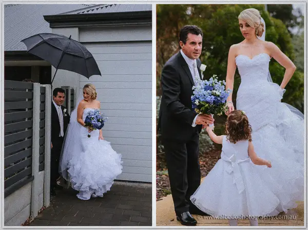 Sarah's father shields her from rain under a black umbrella by a grey weatherboard gate, then hands her off