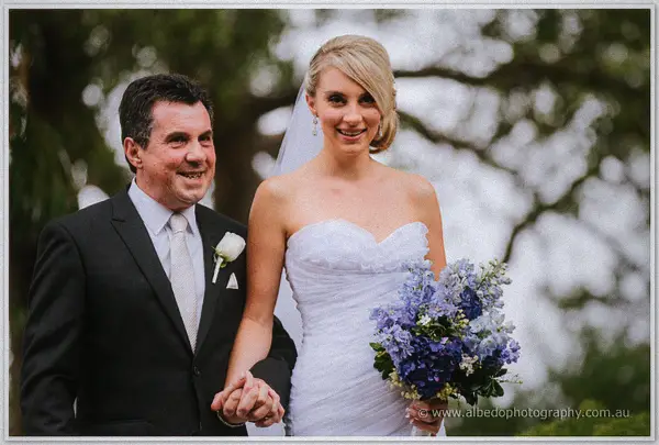 Sarah beams beside her father beneath spreading trees, his hand clasped in hers in rain-soft light