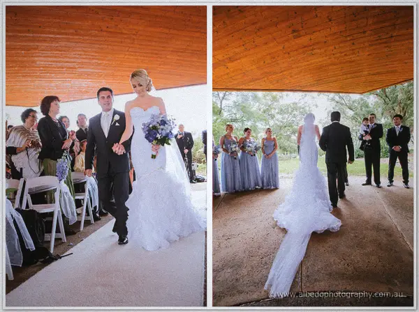 Father and bride walk the white aisle beneath the timber ceiling of the Wildflower Pavilion as guests rise