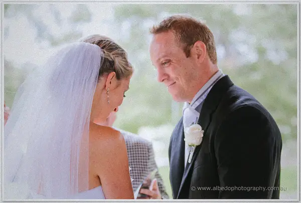 Nathan smiles down at Sarah beneath her veil, a white rose pinned to his lapel, soft trees behind