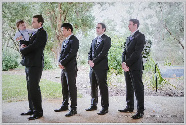 Four groomsmen in black suits wait in line, one bouncing a toddler in braces, under Kings Park gums