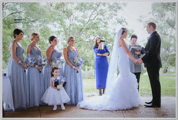 Sarah and Nathan join hands before the celebrant as bridesmaids, flower girl and guests look on