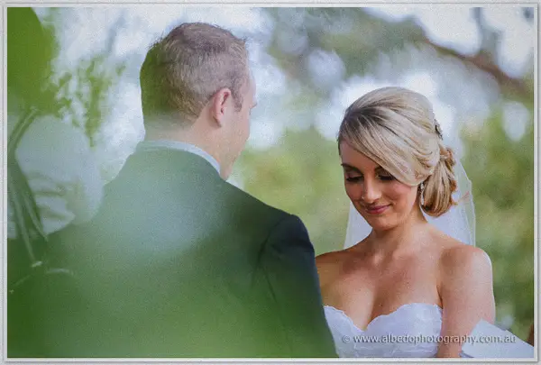 Over Nathan's shoulder, Sarah reads her vows framed by soft green foliage