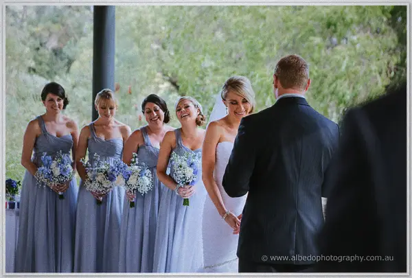Bridesmaids clutching blue and white bouquets beam as Sarah and Nathan face each other to wed
