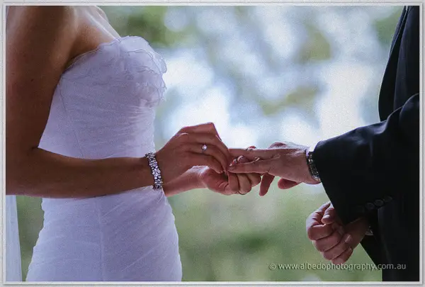 Sarah's bracelet-glittered hand slides a band onto Nathan's finger, green lawns washed out behind