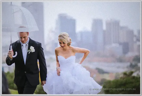 Nathan shields Sarah with an umbrella as wind whips her skirt against the Perth city skyline