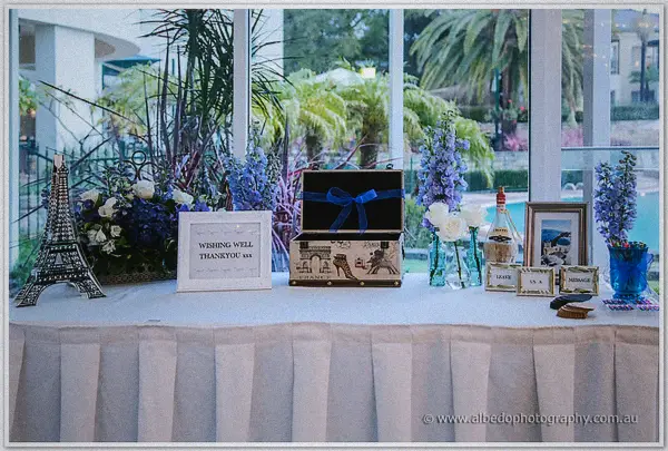 A Paris-themed wishing well table with suitcase card box and blue delphiniums beside the pool