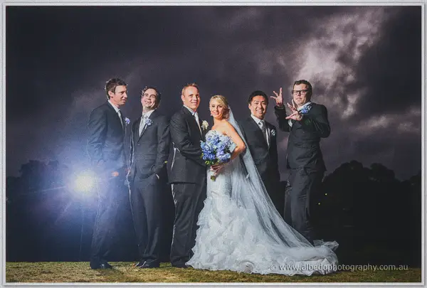 Flash-lit against a brooding night sky, Sarah and Nathan pose with groomsmen, one throwing rock horns