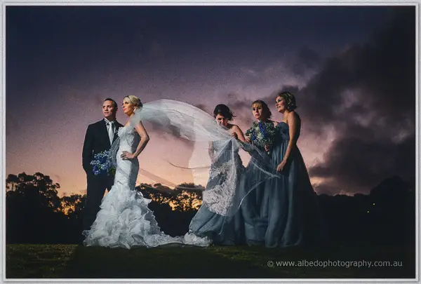 Bridesmaids wrestle Sarah's wind-caught veil at twilight as Nathan watches from her side