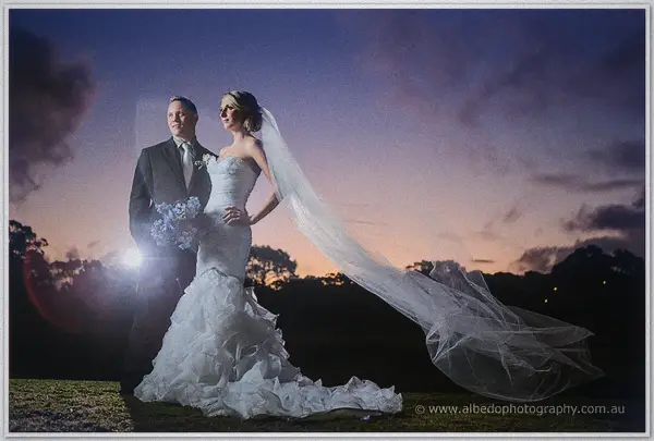 Sarah and Nathan stand on a dusk lawn as wind whips her long veil sideways beneath a purple sunset sky