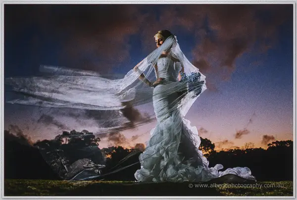 Veil streaming sideways in the evening wind, Sarah grips her bouquet under a violet dusk