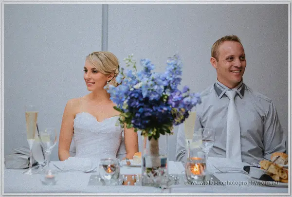 Sarah and Nathan beam from the bridal table behind a tall delphinium centerpiece at Joondalup Resort