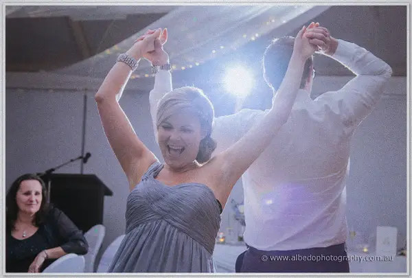 A bridesmaid in grey throws her arms up mid-dance as a flash flares behind her