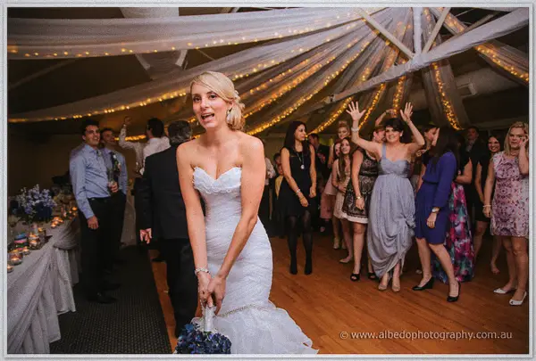 Sarah turns from the crowd with bouquet in hand as guests raise their arms behind her