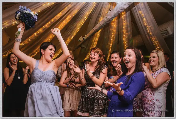 The bouquet catcher raises it in triumph as laughing guests crowd around her
