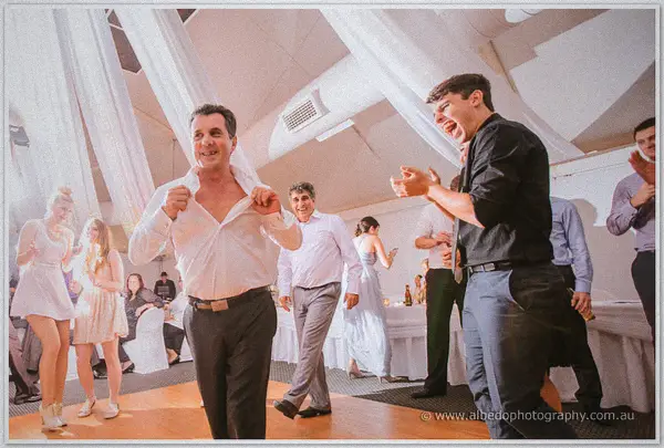 An older guest dances shirt-unbuttoned and fists flying as a younger friend laughs and points