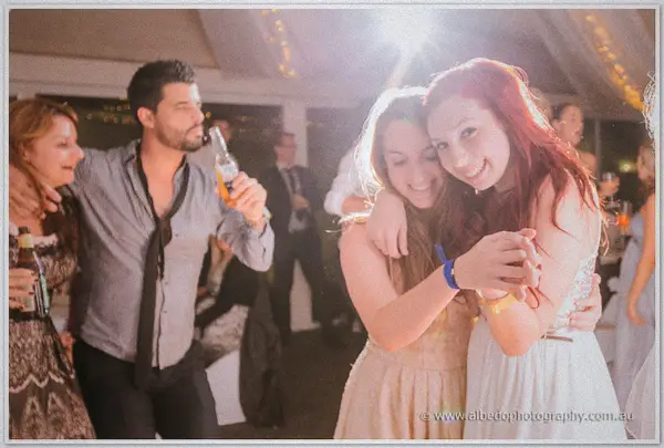 Two friends hug tight on the dance floor, the redhead grinning past a beer-sipping guest