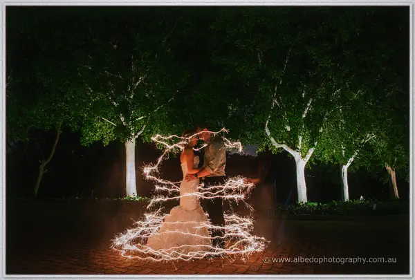 Sarah and Nathan kiss inside swirling rings of sparkler light beneath the uplit trees at night