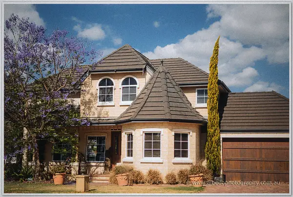 A two-storey cream brick home with tiled turrets sits behind jacaranda blossoms on a bright morning