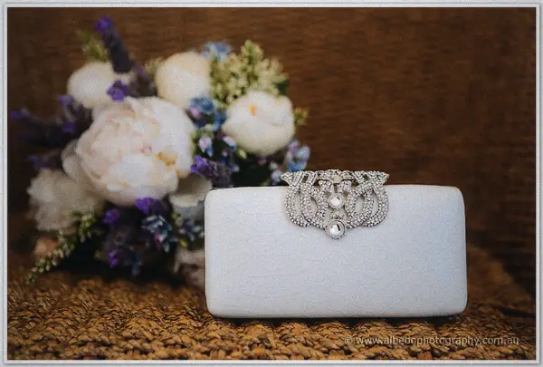 A white satin clutch with a crystal brooch clasp sits before the peony bouquet on textured wicker