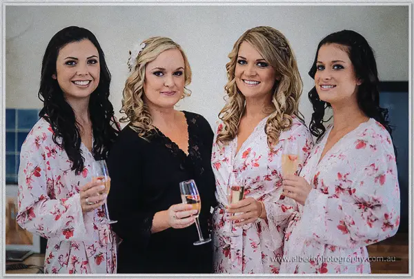 Ashley and three bridesmaids in floral robes raise champagne flutes and smile for the camera indoors