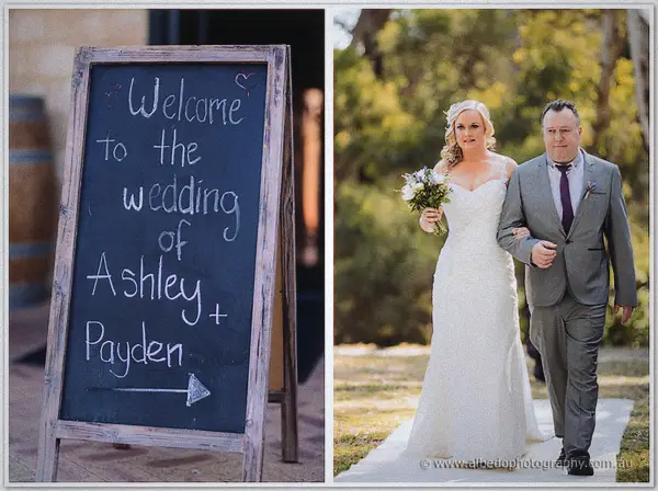 A chalkboard welcome sign stands by wine barrels as Ashley walks the aisle arm in arm with her father