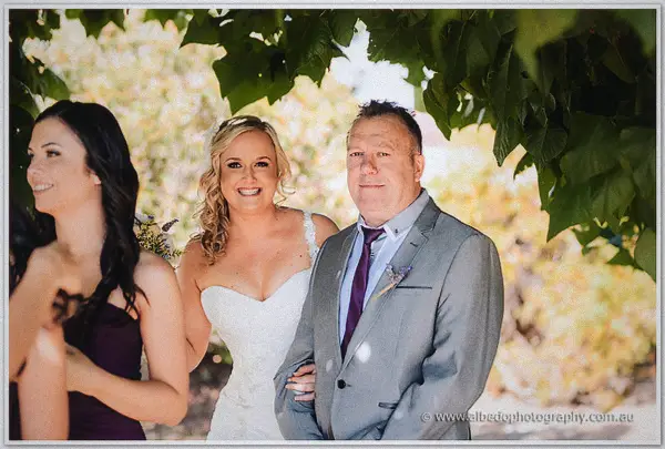 Ashley beams under leafy vines as her father guides her toward the vineyard ceremony