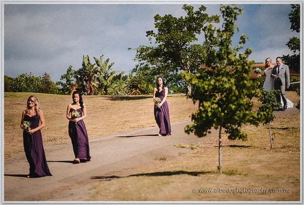 Bridesmaids in plum gowns walk the garden path ahead of Ashley and her father past banana palms