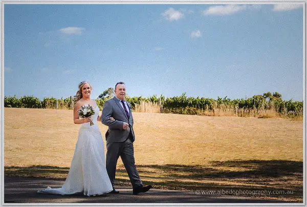 Ashley and her father cross a sunlit paddock track with golden vines stretching behind them