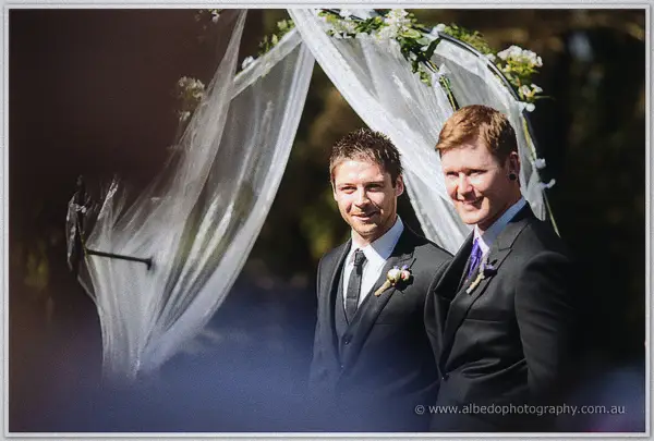 Groom and groomsman smiling beneath a white chiffon-draped wedding arch in bright garden light