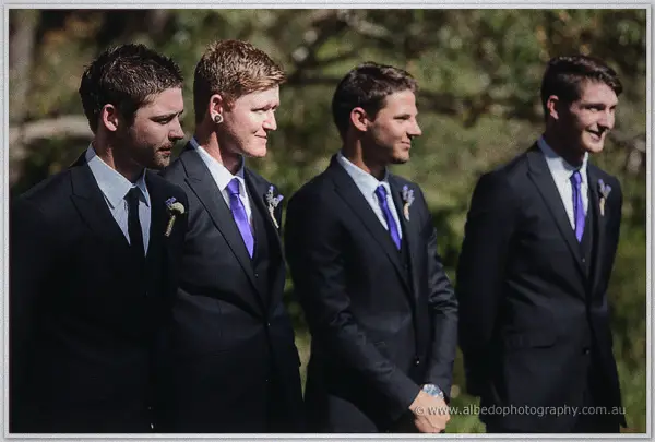 Groomsmen in charcoal suits with purple ties and lavender boutonnieres smile during the ceremony