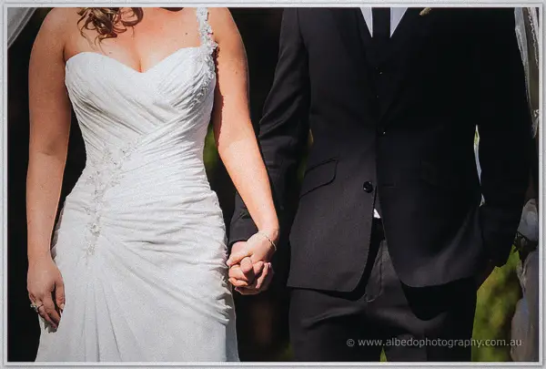 Close-up of the bride and groom holding hands, her ruched gown beside his black suit