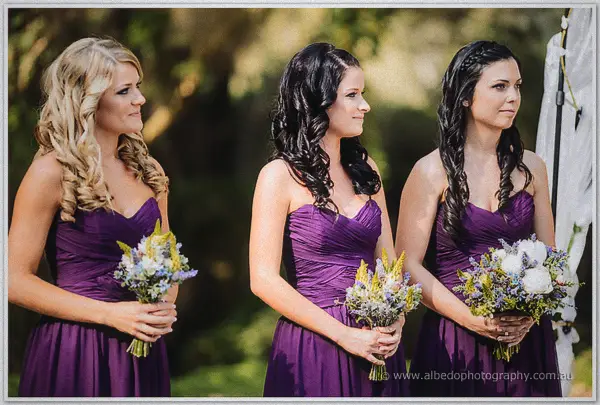 Three bridesmaids in purple gowns holding pastel bouquets at the garden ceremony