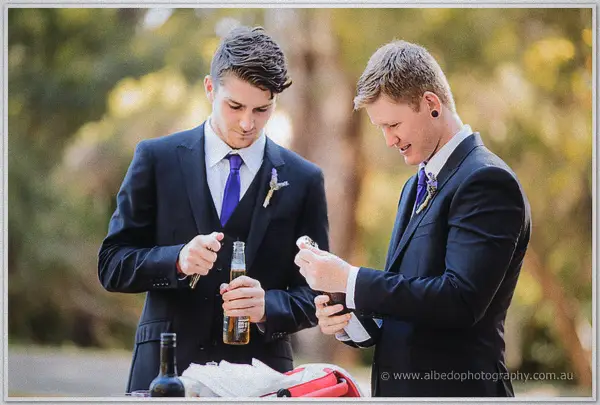 Groomsmen in navy suits and purple ties opening beers among the vines