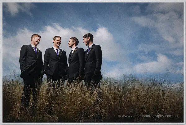 Groomsmen in dark suits laughing among tall golden grasses under a wide blue sky