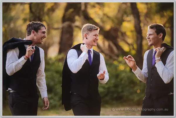 Three groomsmen with purple ties laugh together while carrying their suit jackets through sunlit bushland