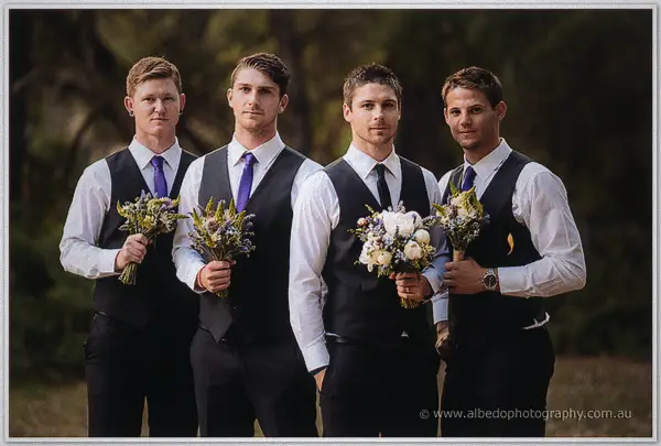 Four groomsmen in black vests and purple ties stand shoulder to shoulder clutching small bouquets