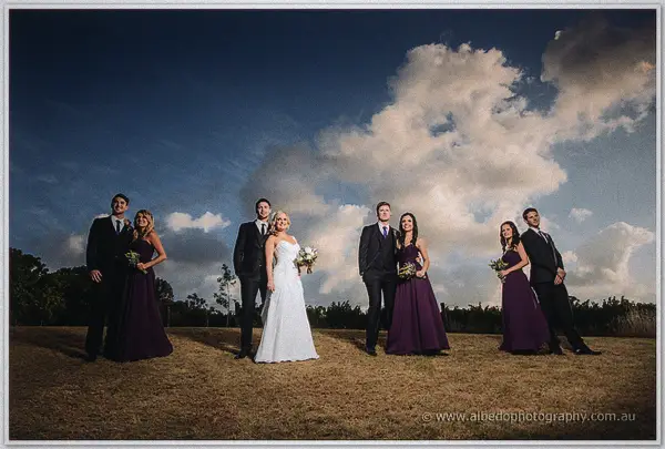 Bridal pairs and groomsmen lean playfully beneath huge blue sky clouds at Carilley Estate