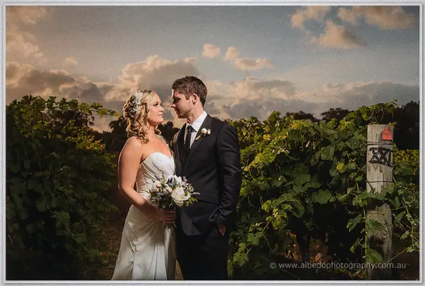 Ashley holds her bouquet beside Payden between leafy vine rows at Carilley Estate under evening cloud