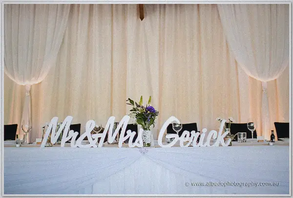 Mr and Mrs Gerick sign stands on the bridal table before draped white curtains and soft light