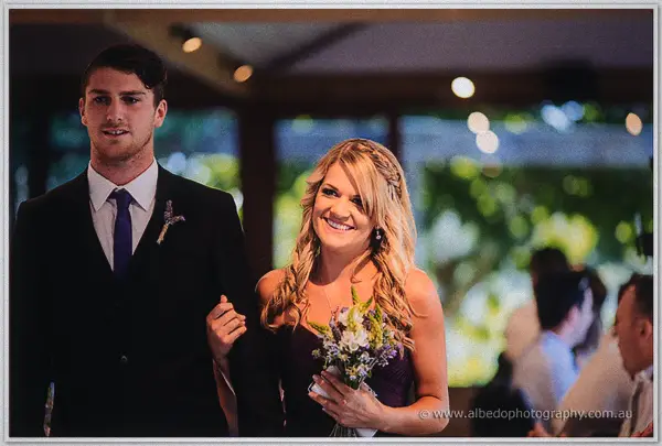 A groomsman escorts a smiling bridesmaid in plum down the aisle, bouquet in hand, guests behind