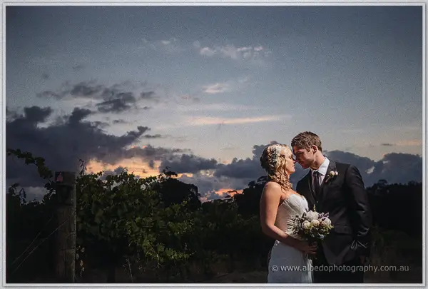 Bride and groom stand close in the vineyard as sunset fires the clouds behind them