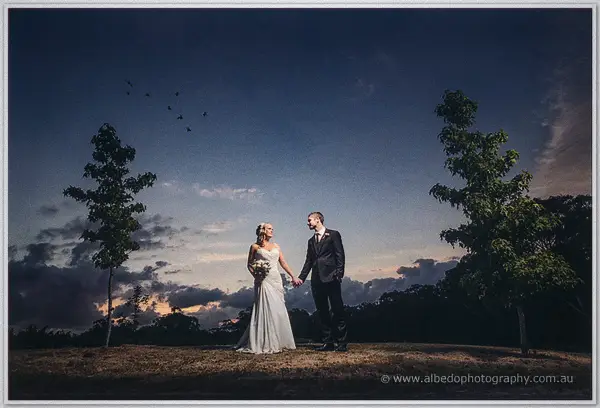 Ashley and Payden hold hands on a paddock hill at dusk beneath scattered clouds and distant birds