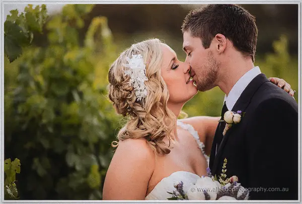 Ashley leans in to kiss Payden close up, her lace hairpiece catching the vineyard's warm glow