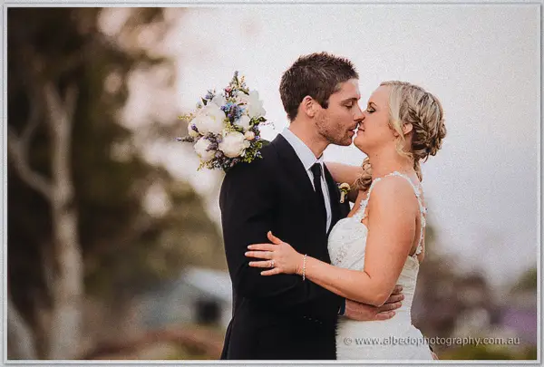 Payden dips to kiss Ashley over her bouquet of white and lilac blooms against soft grey sky
